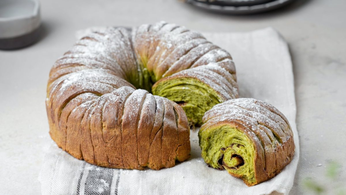 Heller Untergrund. Auf einem hellen Küchentuch ein Matcha-Brot mit Schokolade in Kringelform.