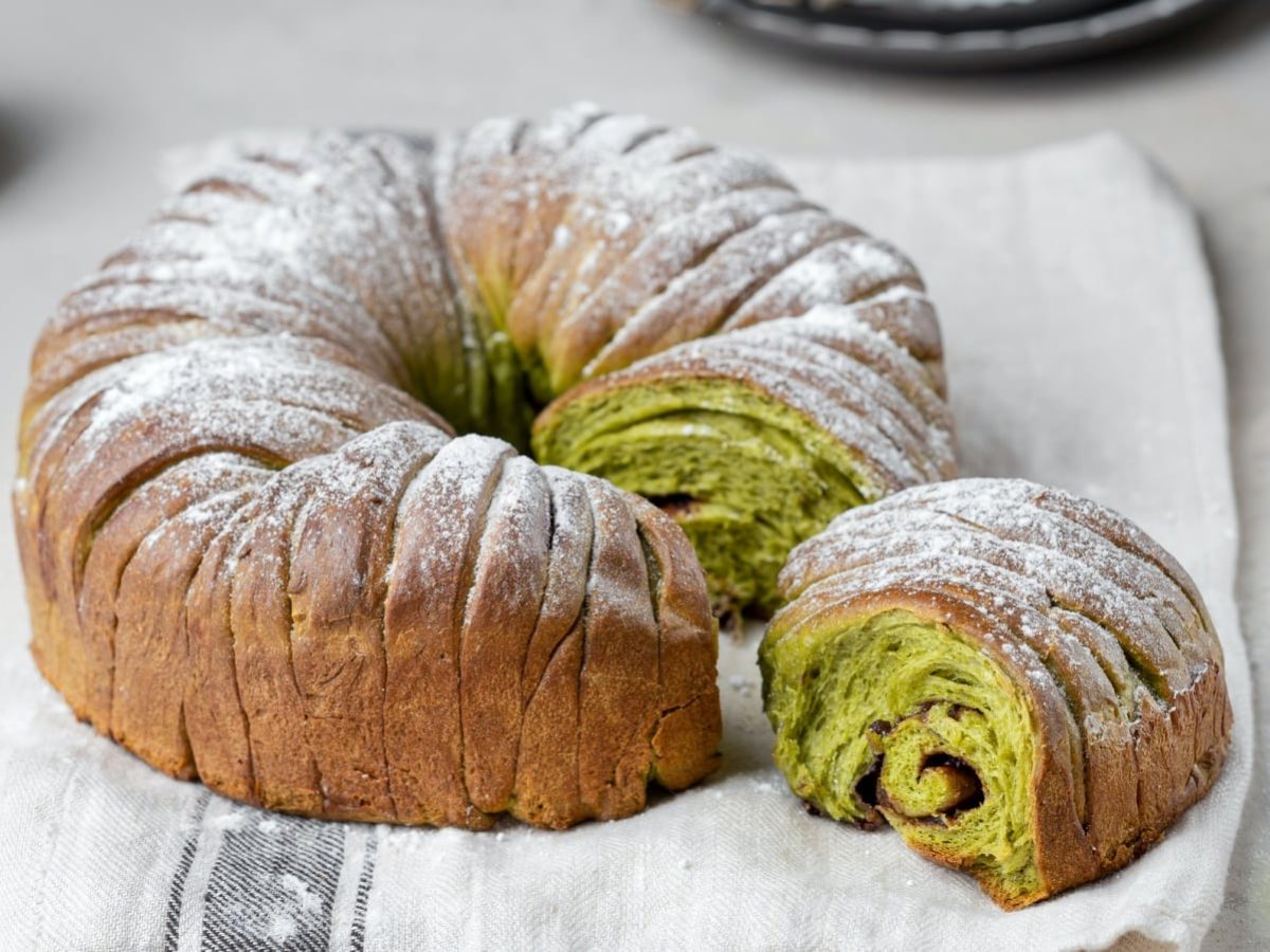 Heller Untergrund. Auf einem hellen Küchentuch ein Matcha-Brot mit Schokolade in Kringelform.