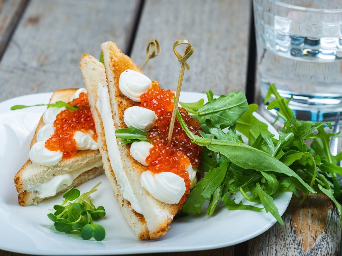 Frontalsicht: Auf einem Holztisch steht einen weißer Teller. Auf dem Teller lieben zwei Mozzarella-Toast mit Forellenkaviar die rechteckig zugeschnitten sind.