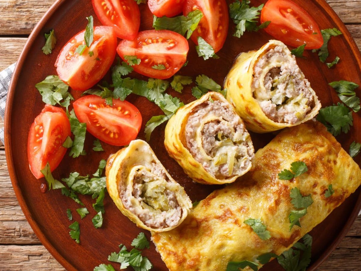 Angeschnittene Omelett-Röllchen mit Hackfleisch-Gurken-Käse-Füllung. Daneben Tomaten.