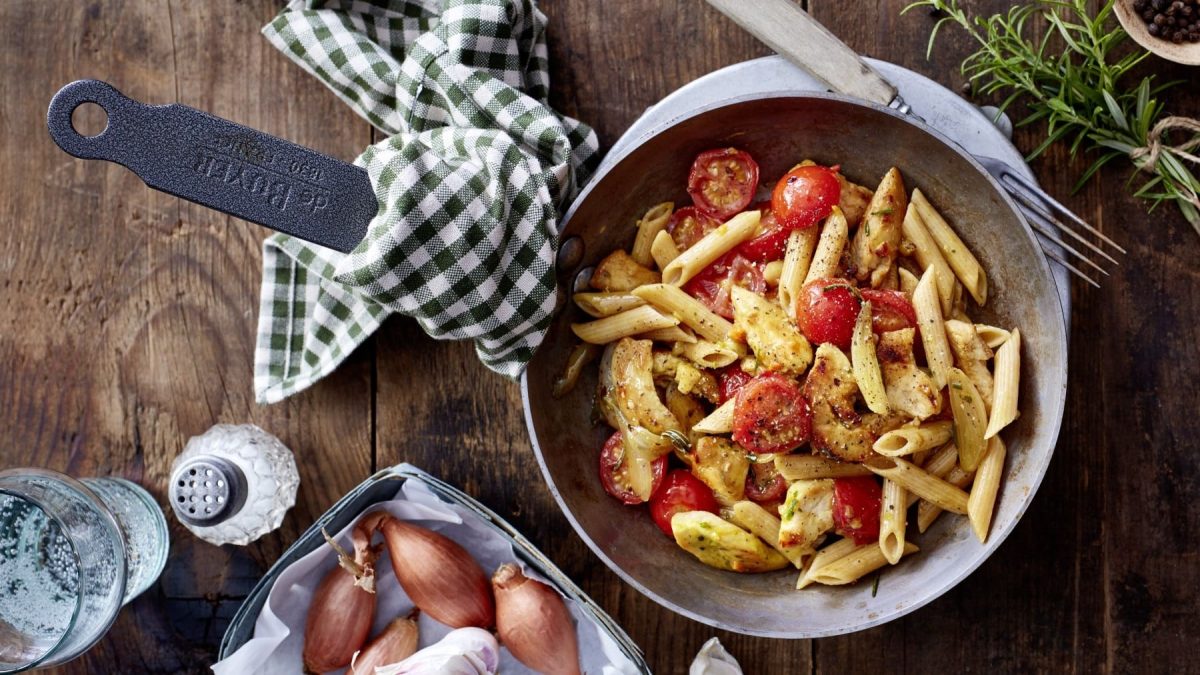 Pasta-Pfanne mit Hähnchen, Cocktailtomaten und Kräutern, serviert in einer großen Pfanne auf einem rustikal-gedeckten Holztisch.