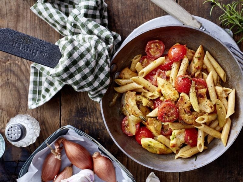 Pasta-Pfanne mit Hähnchen, Cocktailtomaten und Kräutern, serviert in einer großen Pfanne auf einem rustikal-gedeckten Holztisch.