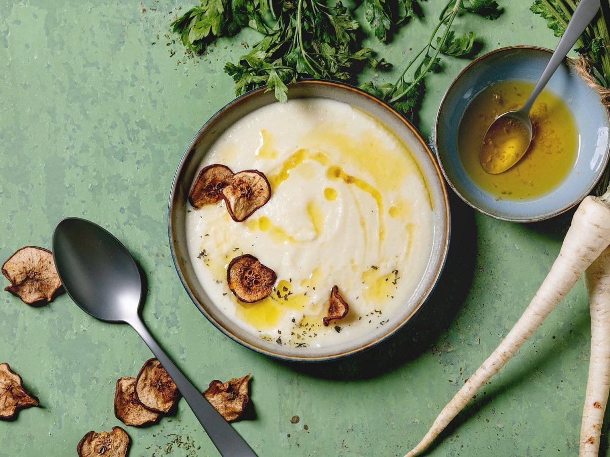 Draufsicht: Auf einem hellen Untergrund steht ein Schale mit Pastinaken-Birnen-Suppe. Drumherum liegen Pastinaken.