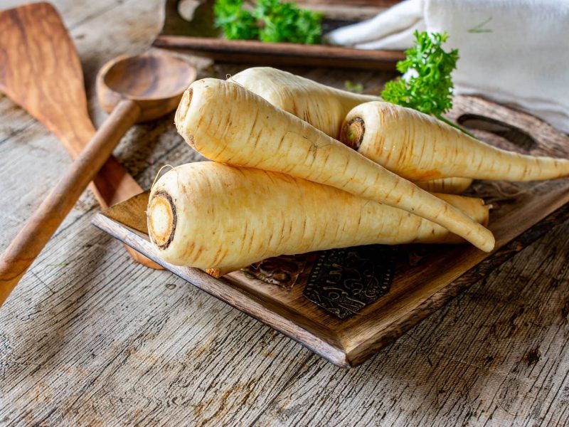 Frontalsicht: auf einem einer holzplatte liegen mehrere unbearbeitete rohe Pastinaken, frische Kräuter und zwei Kochlöffel aus Holz.