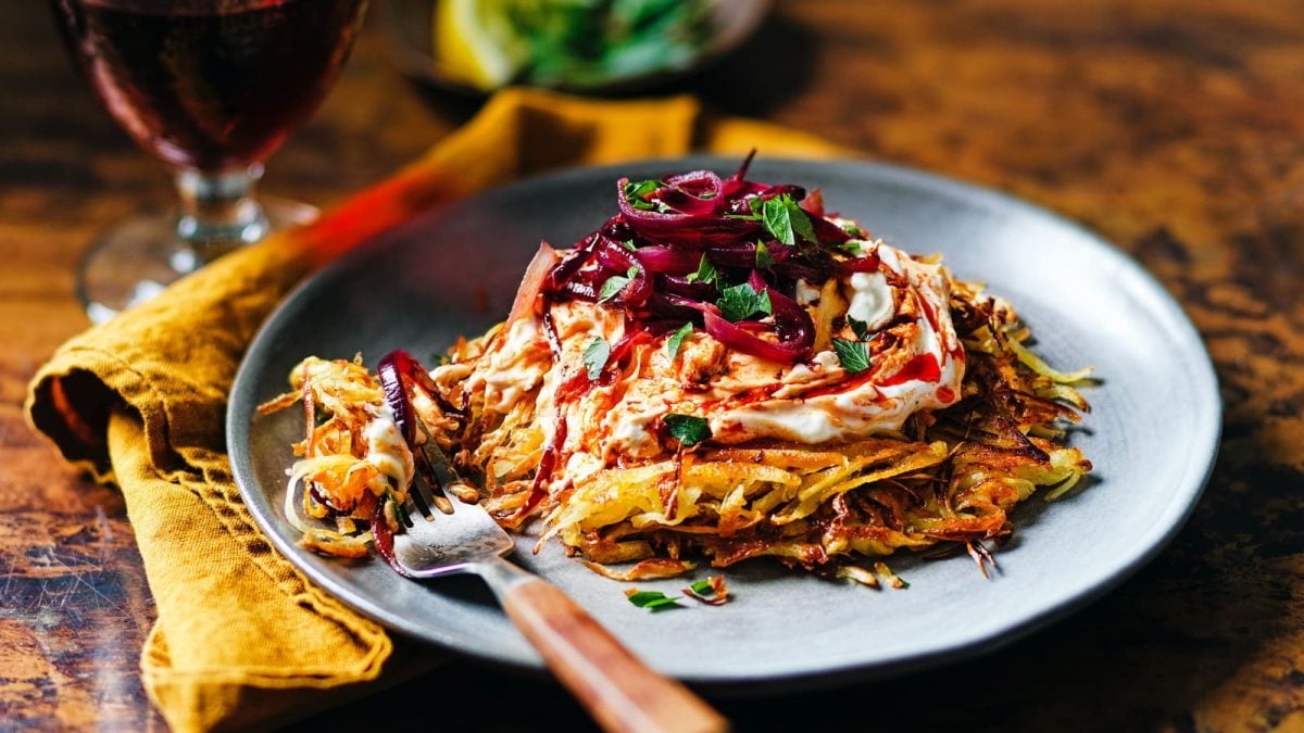 Schrägsicht: Brauner Untergrund auf dem im Bildzentrum ein grauer Teller mit einem Pastinakenrösti mit Harissa-Feta-Creme, karamellisierten Zwiebeln und frischer Petersilie zu sehen ist. Außerdem eine senfgelbe Stoffservierte und eine Gabel mit Holzgriff. Im Hintergrund ein Glas Rotwein.