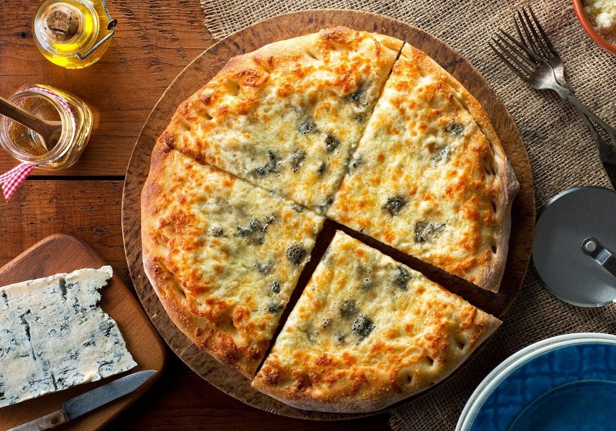 Draufsicht: eine Pizza Margherita mit Blauschimmelkäse die in 4 teile geschnitten wurde liegt auf einem Holztisch. Daneben liegt ein Blauschimmelkäse sowie ein Besteck.