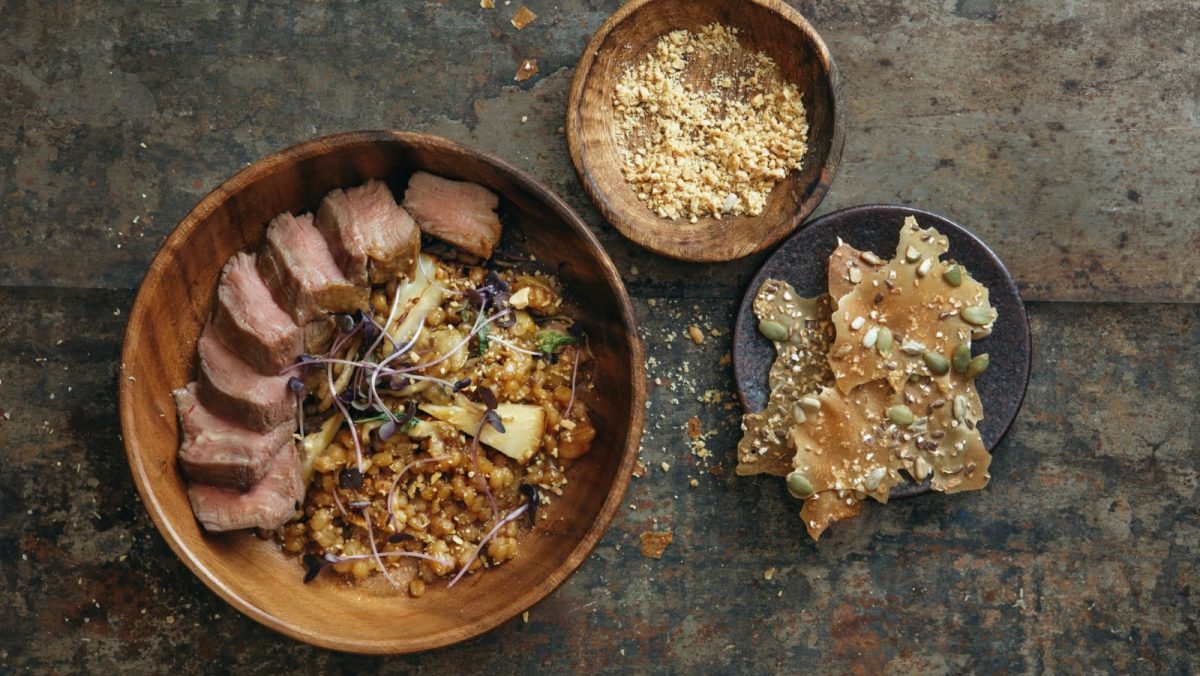 Draufsicht: Dunkler Untergrund mit drei Schalen. Eine mit rustikalem Knäckebrot, eine weitere mit gerösteten Nüsse und eine weitere mit einem aufgeschnittenen Rindersteak mit Getreidesalat, Steinpilzen und Selleriewurzel.