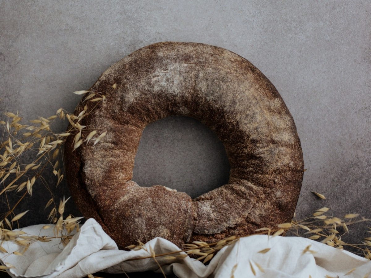 Graue Wand als Hintergrund. Daran lehnt ein Kringel Roggenbrot. Im Bildvordergrund ist außerdem eine weiße Leinenservierte und etwas Hafer zu sehen.