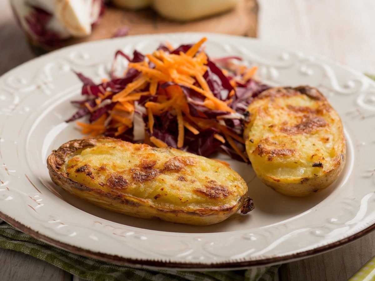 Schräge ansicht: Auf einem Holzuntergrund liegt ein Teller. Auf dem Teller liegen zwei Kartoffelhälften, die mit Käse überbacken wurden. Auch ein Rohkostsalat liegt auf dem teller. Und neben eine eine Gabel.