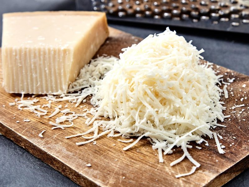 Frontalsicht: Auf einem Brettechen liegt ein stück Parmesa, sowie gerwieber Parmesan. Im Hintergrund ist eine Reibe aus Metall.