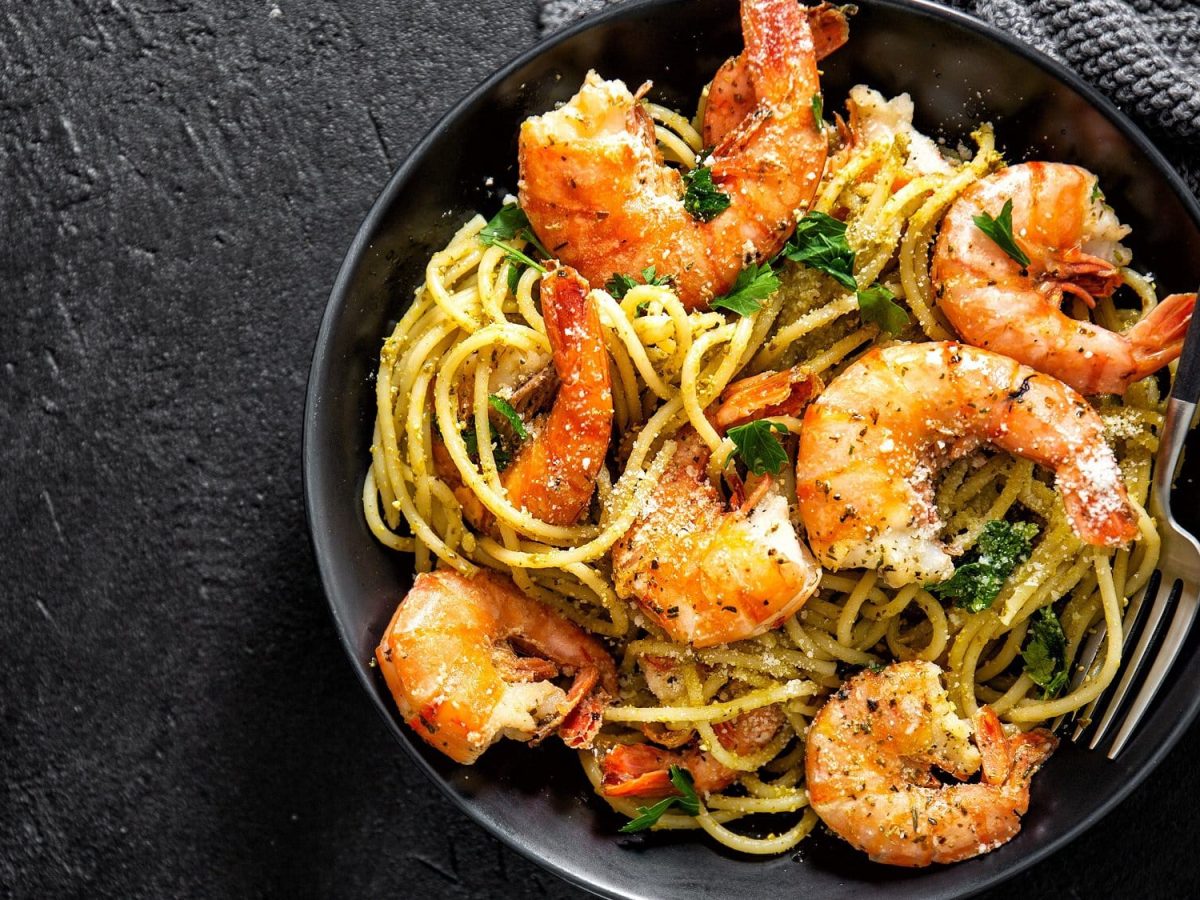 Draufblick; Auf einem dunklen Untergrund liegt eine schwarze Pfanne. In der Pasta mit Pesto und Crevetten und Petersilie.