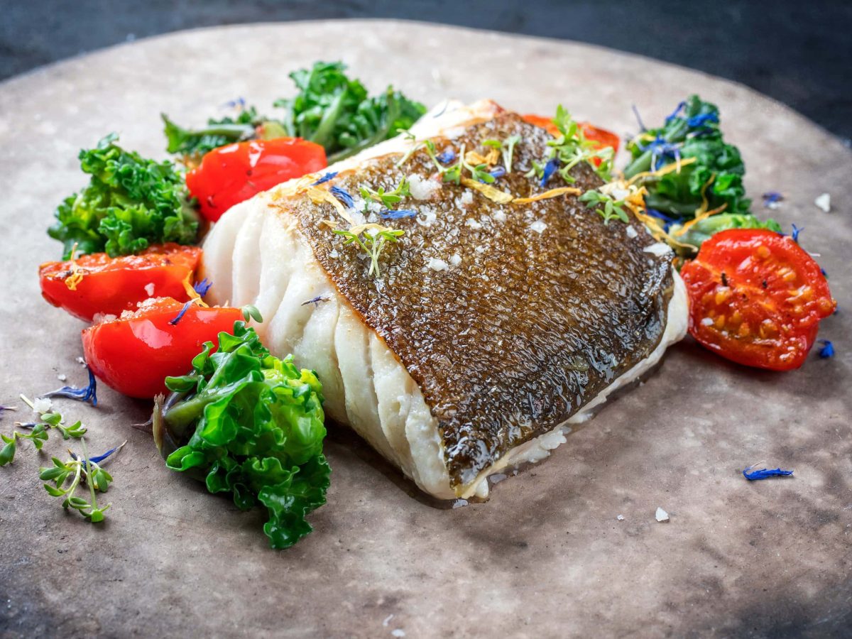 Gebratenes Skrei Loins mit Ofen-Tomaten, Sardellen und Kalettes auf grauem Steinteller. Draufsicht.