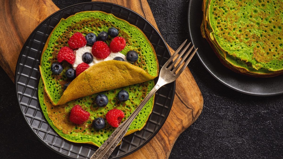 Spinat-Crêpes mit Ricotta und Beeren auf einem schwarzen Teller mit Gabel, darunter ein Holzbrett auf dunklem Hintergrund.