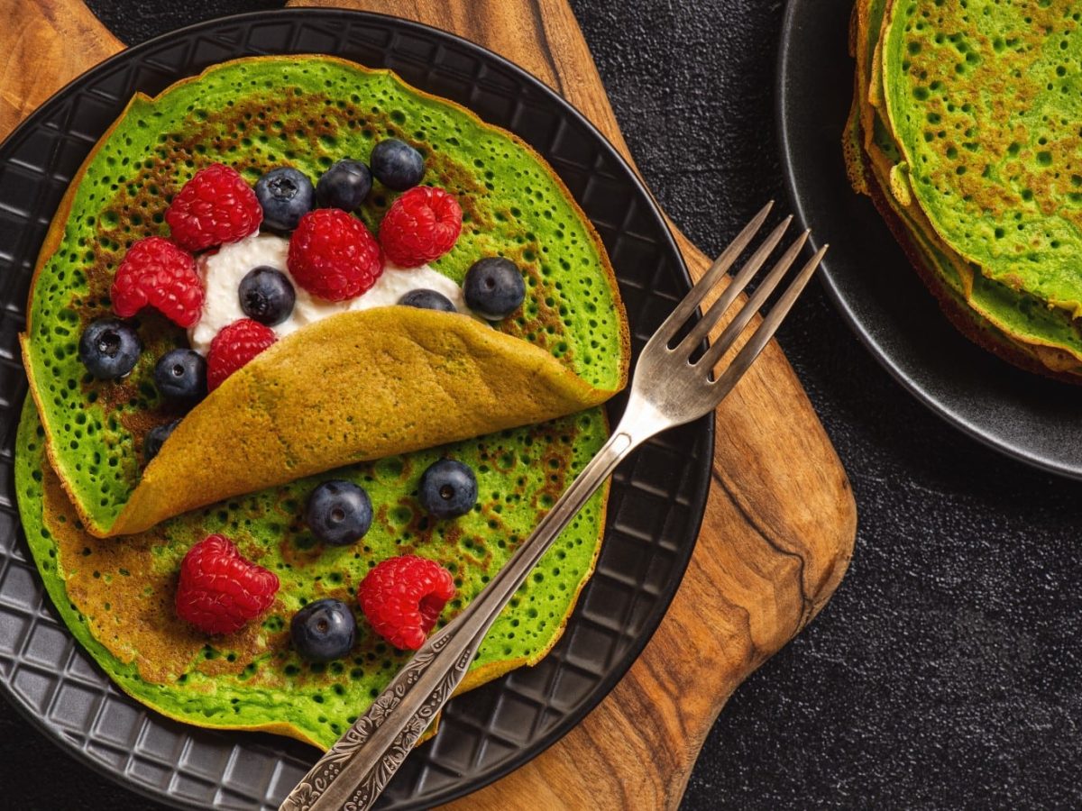 Spinat-Crêpes mit Ricotta und Beeren auf einem schwarzen Teller mit Gabel, darunter ein Holzbrett auf dunklem Hintergrund.