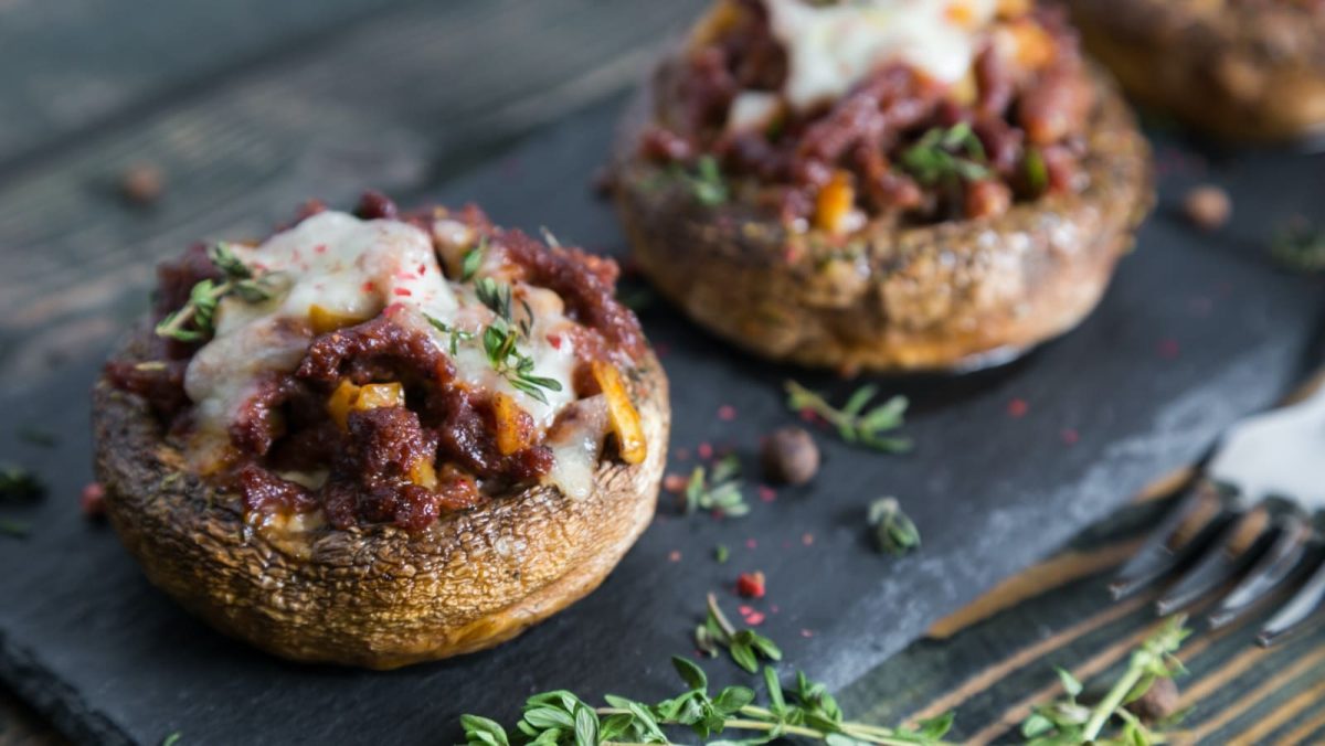 Dunkler Holztisch auf dem eine Schieferplatte liegt auf der stuffed Portobellos angerichtet sind. Davor die Zacken einer Gabel zu sehen und frischer Thymian und roter Pfeffer.