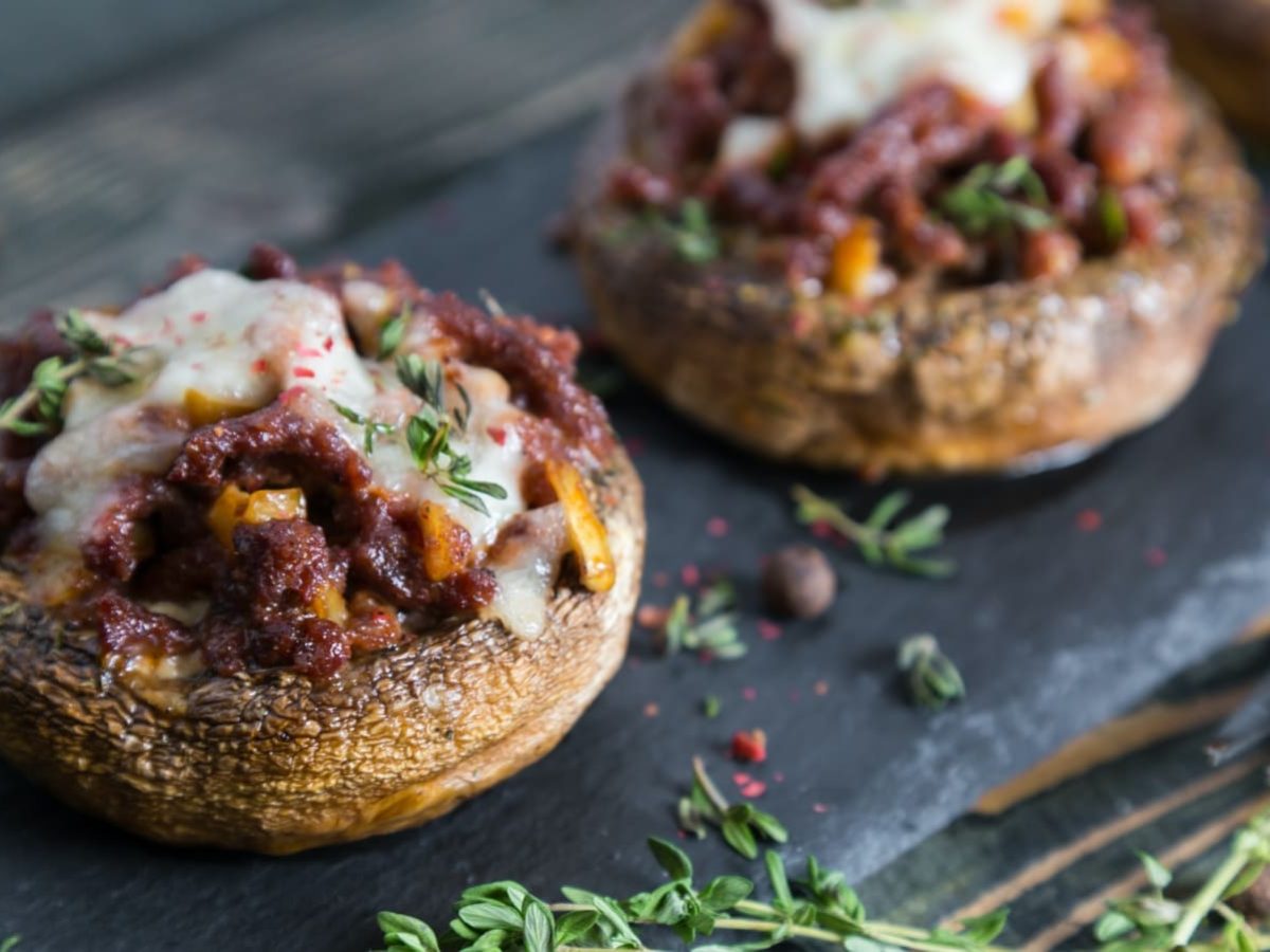 Dunkler Holztisch auf dem eine Schieferplatte liegt auf der stuffed Portobellos angerichtet sind. Davor die Zacken einer Gabel zu sehen und frischer Thymian und roter Pfeffer.