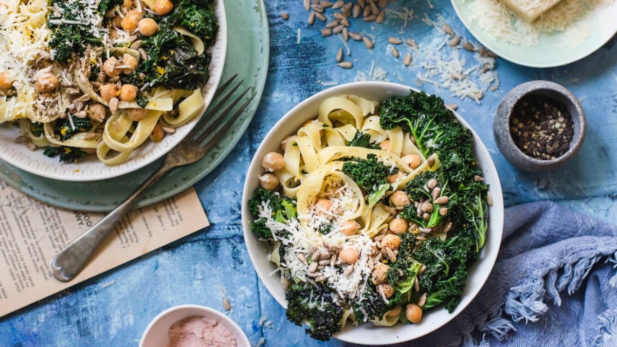 Draufsicht: Blauer Untergrund auf dem zwei Teller, ein Holzbrett, sowie geriebener Parmesan, Sonneblumenkerne und eine Stoffservierte zu sehen sind. Auf den Tellern Tagliatelle mit sautiertem Grünkohl und gerösteten Kichererbsen.