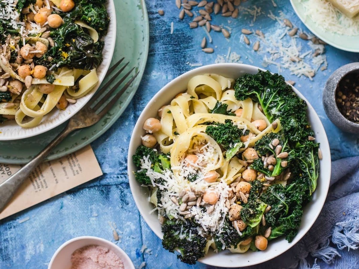 Draufsicht: Blauer Untergrund auf dem zwei Teller, ein Holzbrett, sowie geriebener Parmesan, Sonneblumenkerne und eine Stoffservierte zu sehen sind. Auf den Tellern Tagliatelle mit sautiertem Grünkohl und gerösteten Kichererbsen.