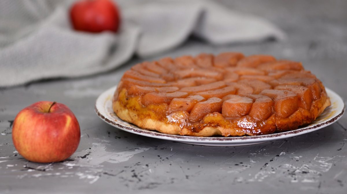 Ein weißer Teller mit der glänzenden Tarte-Tatin auf einem grauen Untergrund mit einem Apfel.