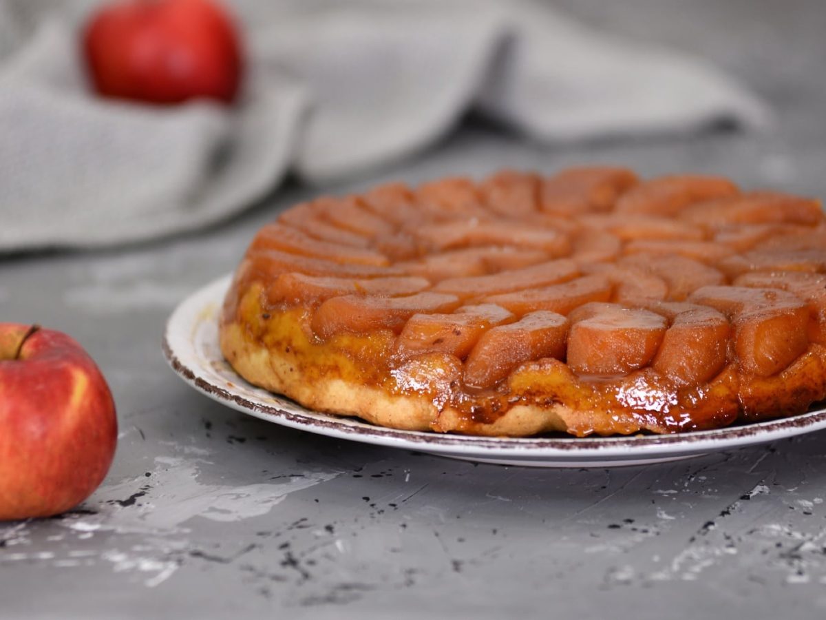 Ein weißer Teller mit der glänzenden Tarte-Tatin auf einem grauen Untergrund mit einem Apfel.