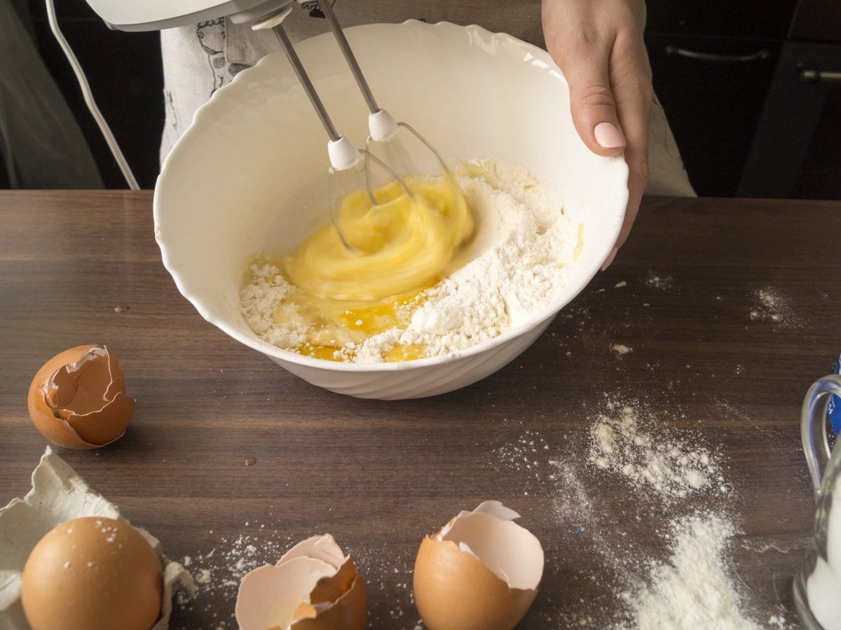 auf einen dunklem Holztisch liegen Mehlreste und aufgeschlagene Eierschalen- Eine hand hält eine weiße Schüssel mit Eiern und hält ein Handrührgerät mit Knethaken in der Hand.