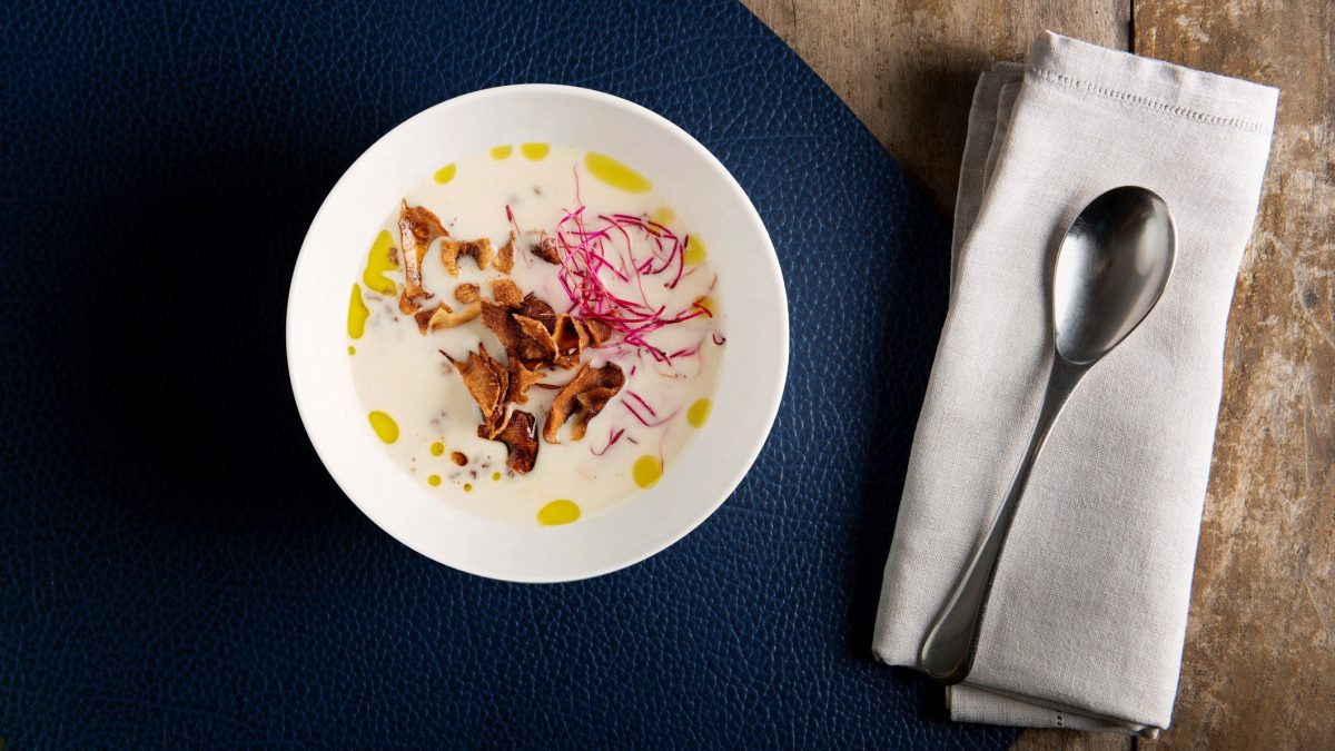 Topinambursüppchen mit Rote Bete, Topinambur-Chips, Speck und Kräuteröl in weißem Suppenteller auf schwarzer Unterlage auf Holztisch. Daneben Suppenlöffel auf Serviette. Draufsicht.