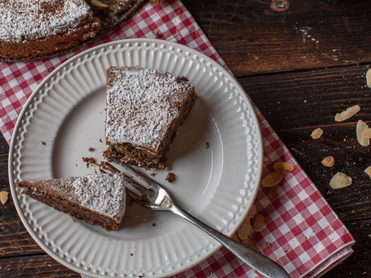 Torta Caprese al cioccolato Stück auf weißem Teller auf rot-weißem Tuch mit Kuchengabel. Daneben Mandelsplitter. Oben Torta Caprese al cioccolato im Ganzen. Draufsicht auf dunklem Holztisch.