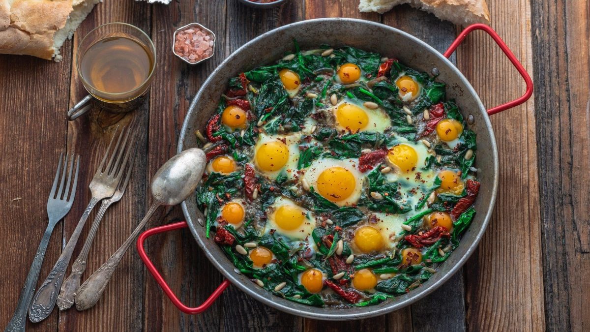 Draufsicht: Auf einem dunklen Holztisch steht eine runde Metallpfanne mit roten Griffen. In der ist das traditionelles türkisches Spinatgericht Ispanak Yumurta. Auf dem Spinat sind Spiegeleier. Drumherum liegen Besteck, Brot und Gewürze.