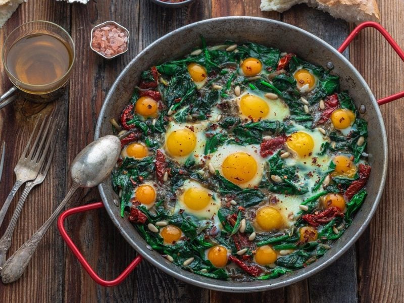 Draufsicht: Auf einem dunklen Holztisch steht eine runde Metallpfanne mit roten Griffen. In der ist das traditionelles türkisches Spinatgericht Ispanak Yumurta. Auf dem Spinat sind Spiegeleier. Drumherum liegen Besteck, Brot und Gewürze.