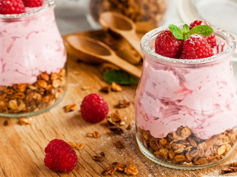 Zwei Gläser mit Himbeer-Mousse und Müsli, dekoriert mit frischen Himbeeren, Minze und Puderzucker. Darum liegen Himbeeren und Müsli.