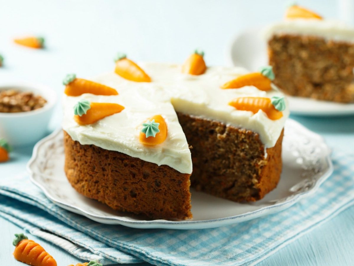 Ein angeschnittener veganer Schoko-Karottenkuchen mit weißem Frosting auf einem Teller, dahinter steht ein weiterer Teller mit einem Stück Kuchen.