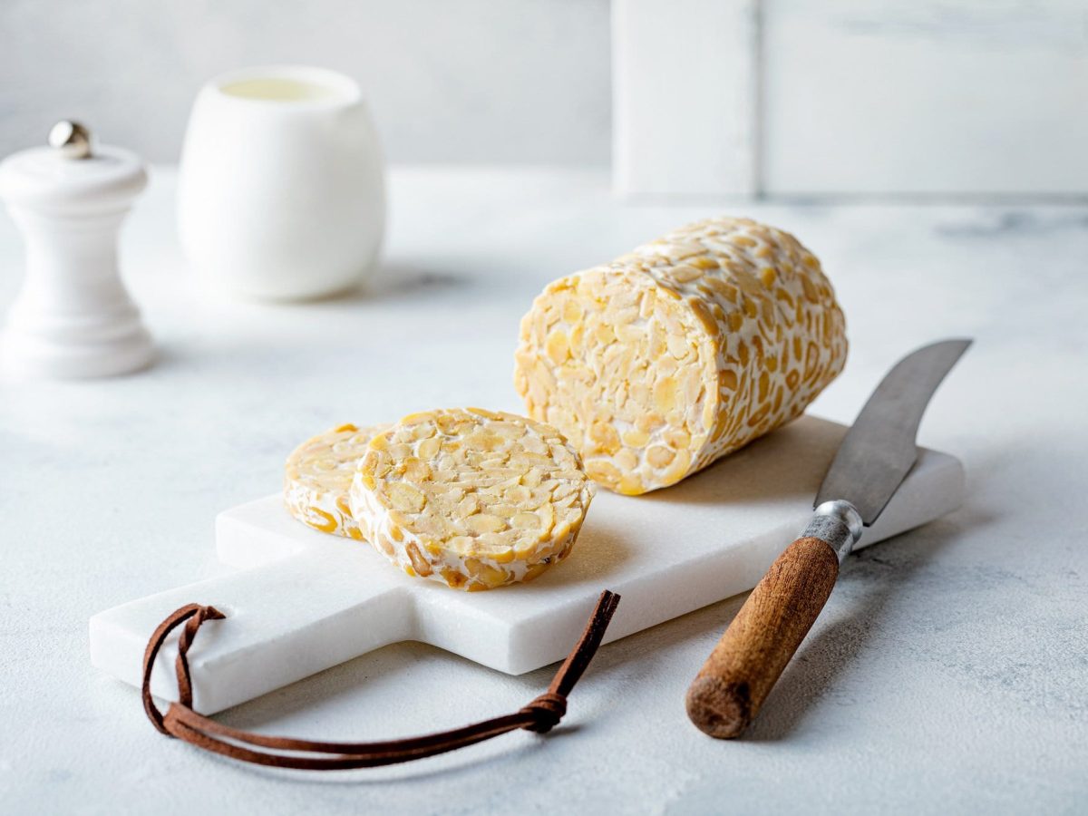 Eine angeschnittene Rolle Tempeh auf einem weißen Keramikbrett, daneben ein Messer, im Hintergrund ein Salzstreuer.