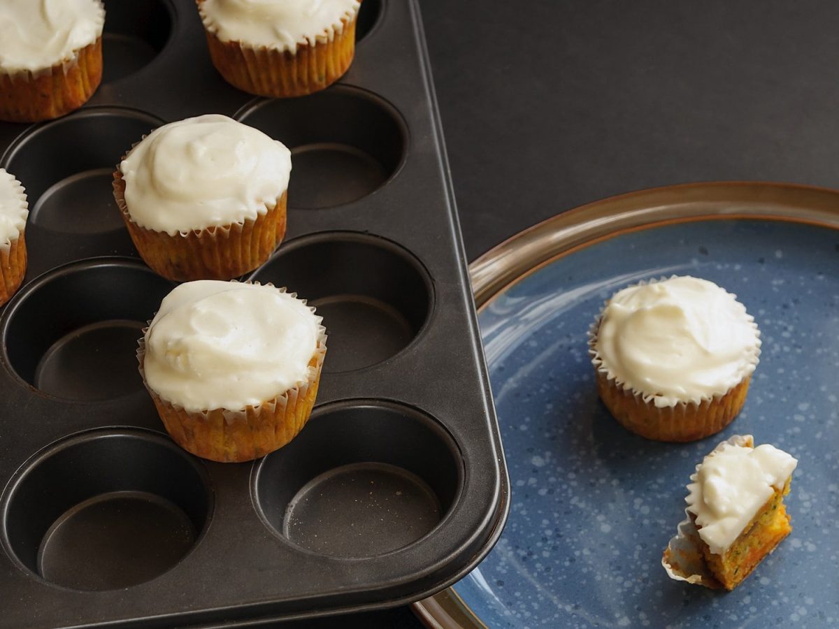 Ein Muffinblech mit Zimtschnecken-Muffins mit Frosting, daneben ein Teller, auf dem weitere Muffins liegen.