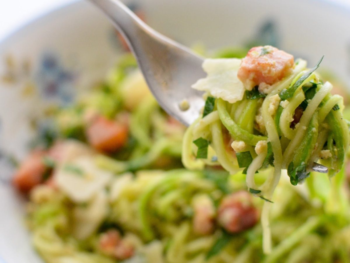 Ein weiß-blauer Teller mit der Zucchini-Carbonara und eine rGAbel mit aufgerollter Zucchini und der Sauce.