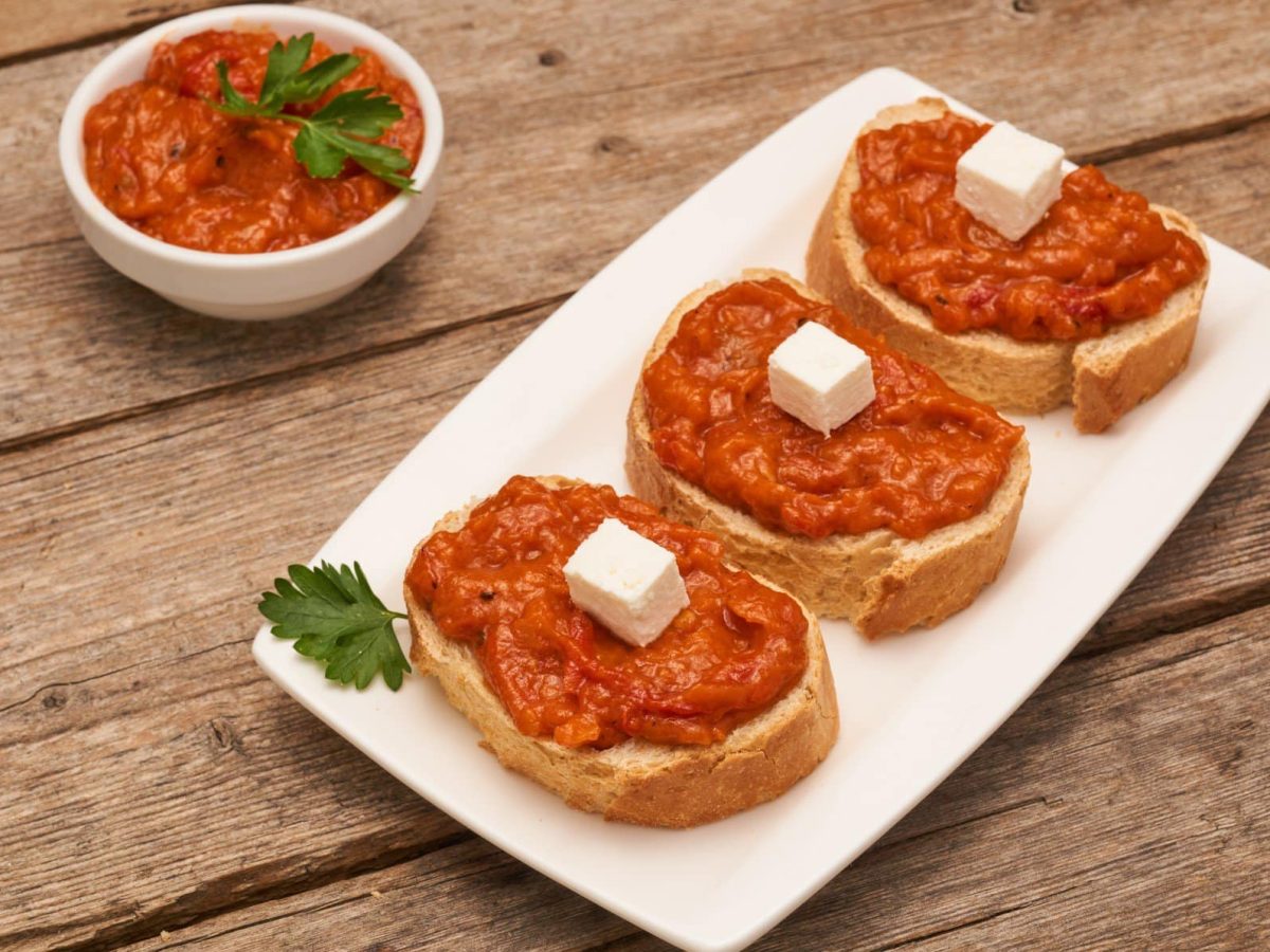 Ajvar-Feta-Creme mit Feta-Würfel auf drei Scheiben Brot auf weißem Teller und Holz. Daneben Schale mit der Creme. Draufsicht.