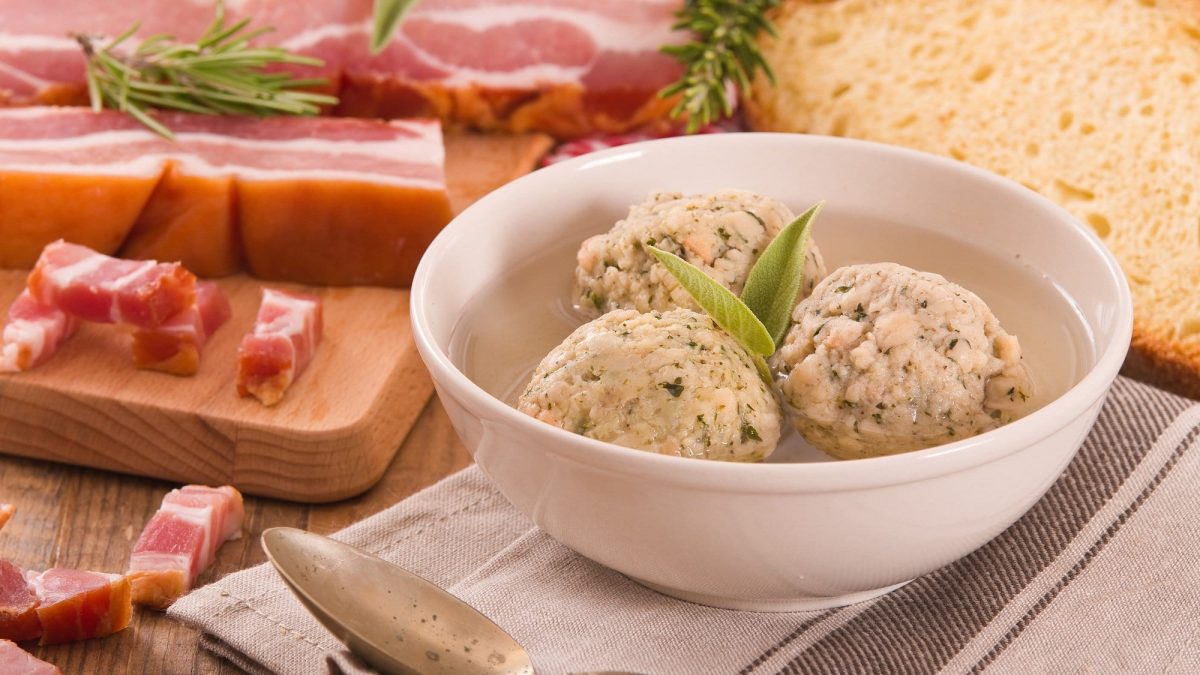 Frontalsicht, schräg, oben: IN einer weißen Schüssel sind drei Südtiroler Buchweizenknödel. Dahinter liegen Speckschwarten und Kräuter.