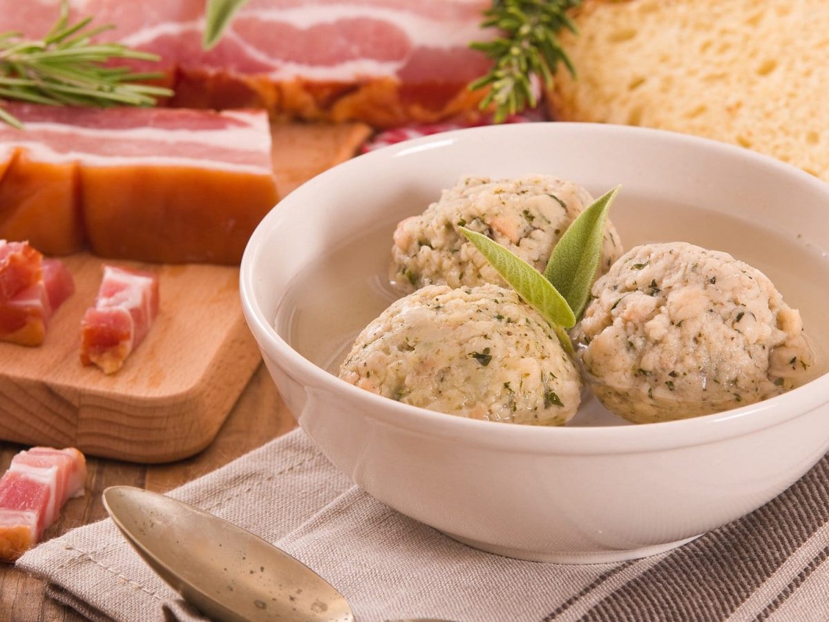 Frontalsicht, schräg, oben: IN einer weißen Schüssel sind drei Südtiroler Buchweizenknödel. Dahinter liegen Speckschwarten und Kräuter.