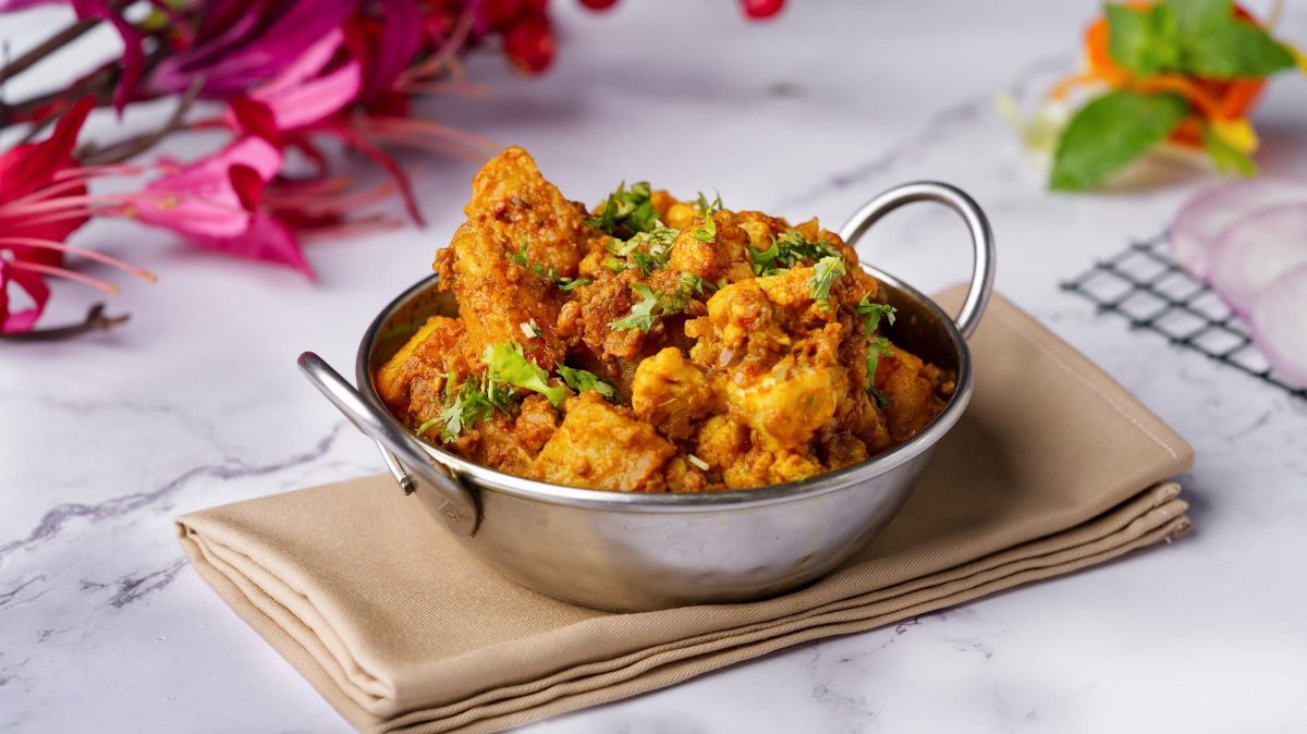 Eine Metallschüssel mi Aloo Gobi, ein Gericht aus Kartoffeln und Blumenkohl. Die Schale steht auf einer Serviette, das Gericht im mit Koriander bestreut.