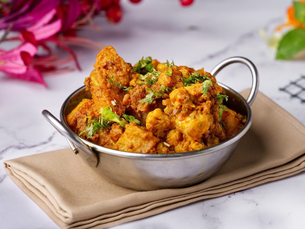 Eine Metallschüssel mi Aloo Gobi, ein Gericht aus Kartoffeln und Blumenkohl. Die Schale steht auf einer Serviette, das Gericht im mit Koriander bestreut.