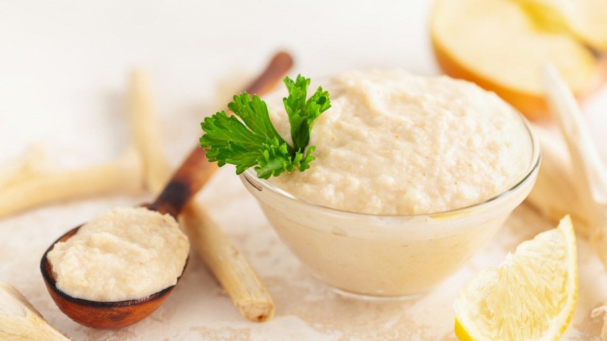 Frontalsicht: Ein Glasschälchen mit einem Apfel-Meerrettich-Mousse steht auf einem hellem Untergrund. Drum herum liegen Meerrettischwurzeln, Zitronenschnitzer und eine Apfelhälfte.