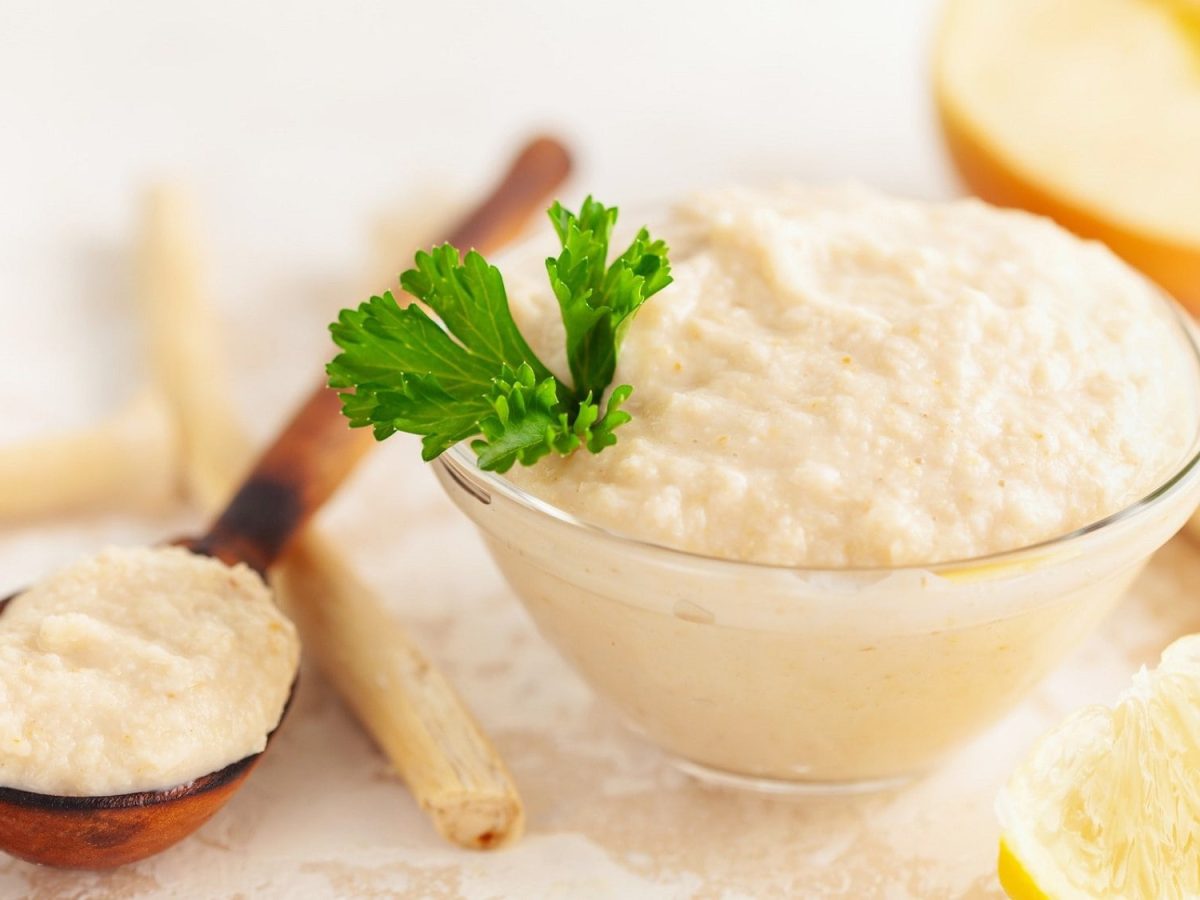 Frontalsicht: Ein Glasschälchen mit einem Apfel-Meerrettich-Mousse steht auf einem hellem Untergrund. Drum herum liegen Meerrettischwurzeln, Zitronenschnitzer und eine Apfelhälfte.