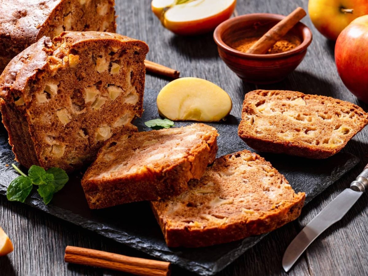 Draufsicht: Ein Apfel-Zimt-Brot liegt aufgeschnitten mit Äpfeln und Zimt auf einem dunkleren Holztisch.