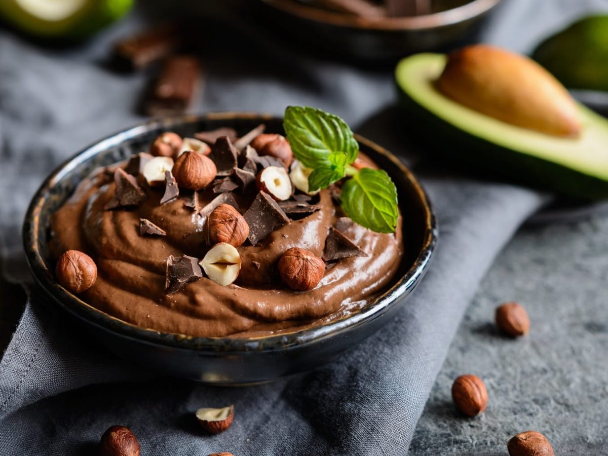 Eine schwarze Schale mit dem Avocado-Schokopudding auf grauem Untergrund. Im Hintergrund unscharf eine Avocado und ein paar Nüssen.