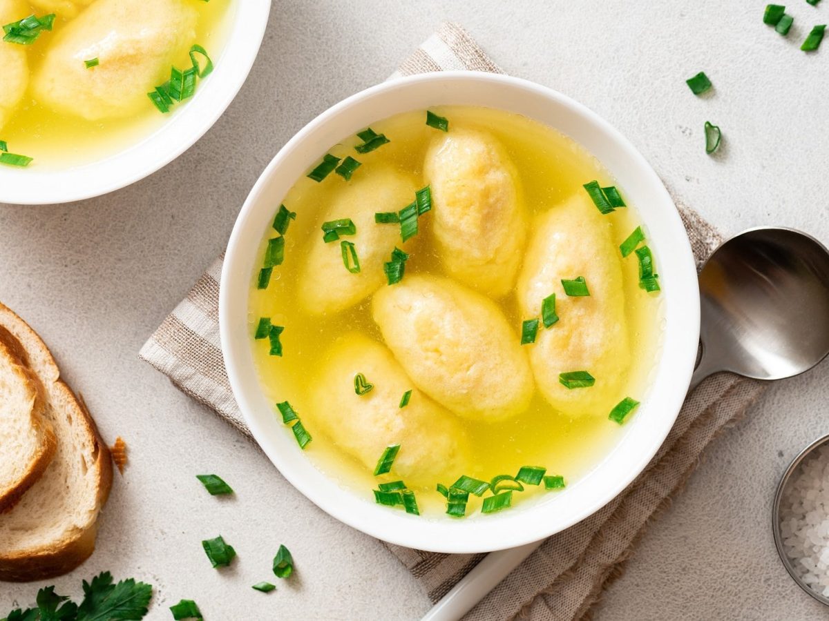 Draufsicht: zwei Schüsseln Bayerische Grießnockerlsuppe. Drumherum stehen ein Salzgefäß, Löffel, Brotscheiben und frische Kräuter.