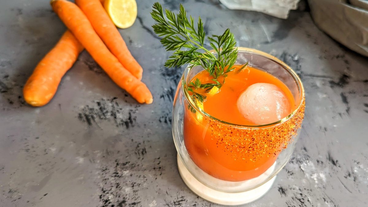 Ein Glas mit dem Bunny Mary auf grauem Untergrund. Daneben Karotten, Zitrone, Eis und ein graues Tuch.