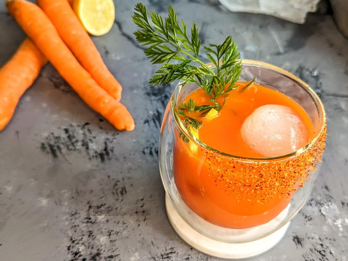 Ein Glas mit dem Bunny Mary auf grauem Untergrund. Daneben Karotten, Zitrone, Eis und ein graues Tuch.