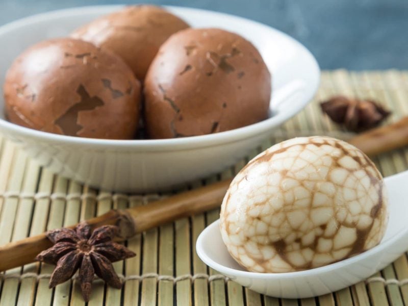 Chinese Tea Eggs chinesische Tee-Eier mit Schale in einer weißen Schüssel auf Bast. Auf weißem Löffel ein geschältes. Frontalansicht.