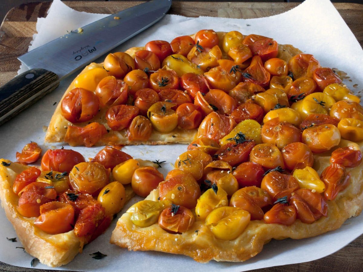 Angeschnittene Cocktailtomaten-Tarte-Tatin auf Backpapier auf dunklem Untergrund. Daneben ein Kuchenmesser. Draufsicht.