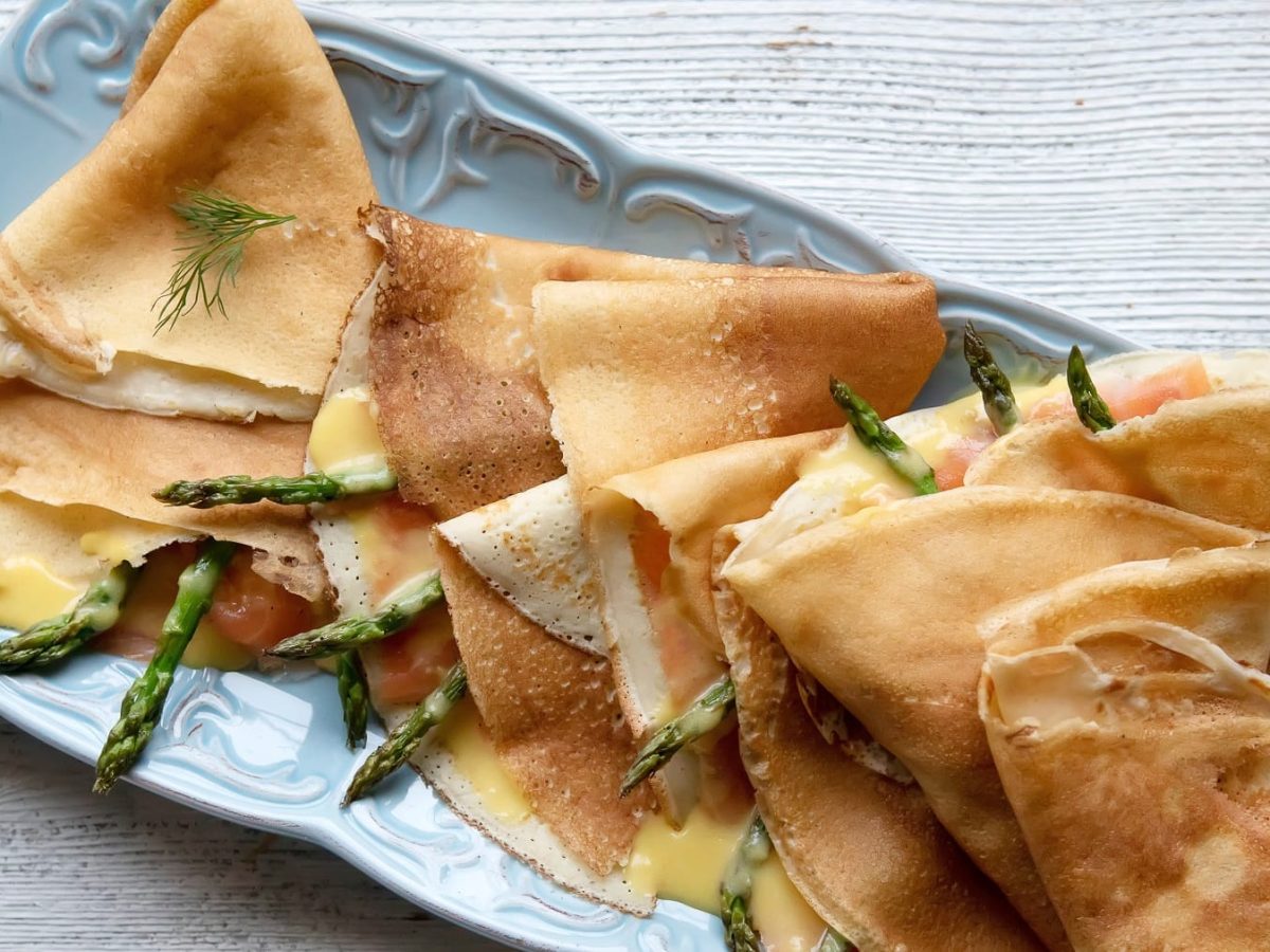 Einige Crepes mit Lachs und Spargel gefüllt und zu Viertel gefalten auf einem blauen länglichen Teller von oben fotografiert.