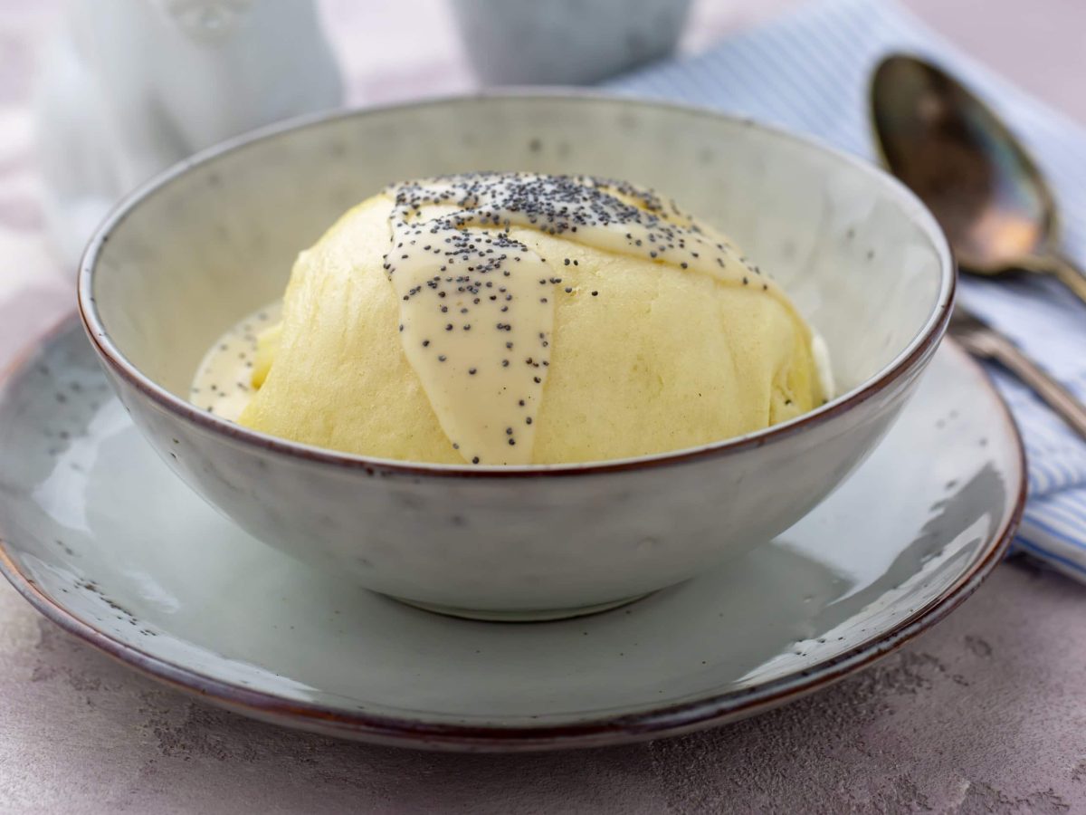 Eine Dampfnudel in einem weißen tiefen Teller auf einem Steintisch mit Löffeln.